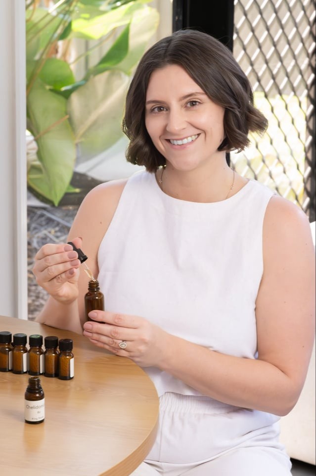 Ash Mastropasqua, homeopath of The Sacred Return. Wearing light, natural / neutral tones made of linen material. Smiling, seated as a desk, holding a homeopathic remedy that she has just made with the equipment on the desk - homeopathic medicine from the Homeopathic pharmacy and amber bottles.