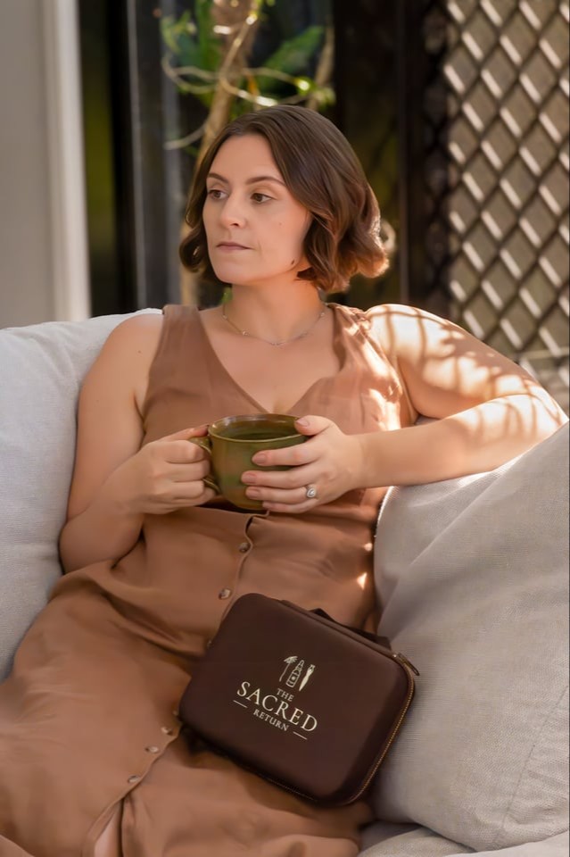 Ash Mastropasqua, homeopath of The Sacred Return. Wearing a chocolate brown dress made of linen material. Ash is sitting comfortably on the couch, looking off to the side deep in thought. Holding a cup of tea and on her lap is the dark brown The Sacred Return Homeopathic First aid Kit case.