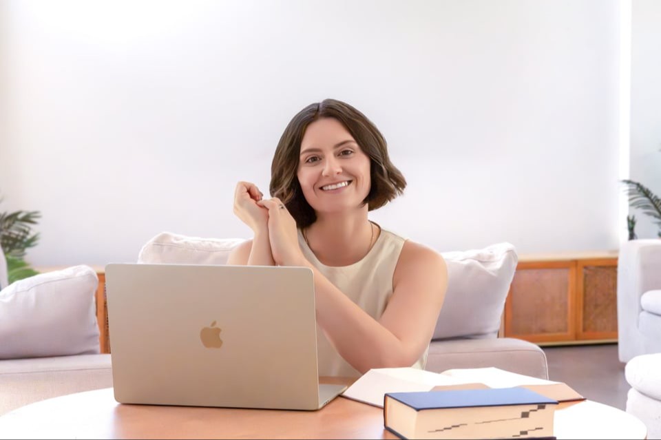 Ash Mastropasqua, homeopath of The Sacred Return. Wearing light, natural / neutral tones made of linen material. Sitting at a desk in front of her computer and homeopathic books on the desk. She is looking up and smiling with her hands clasped to her chin.