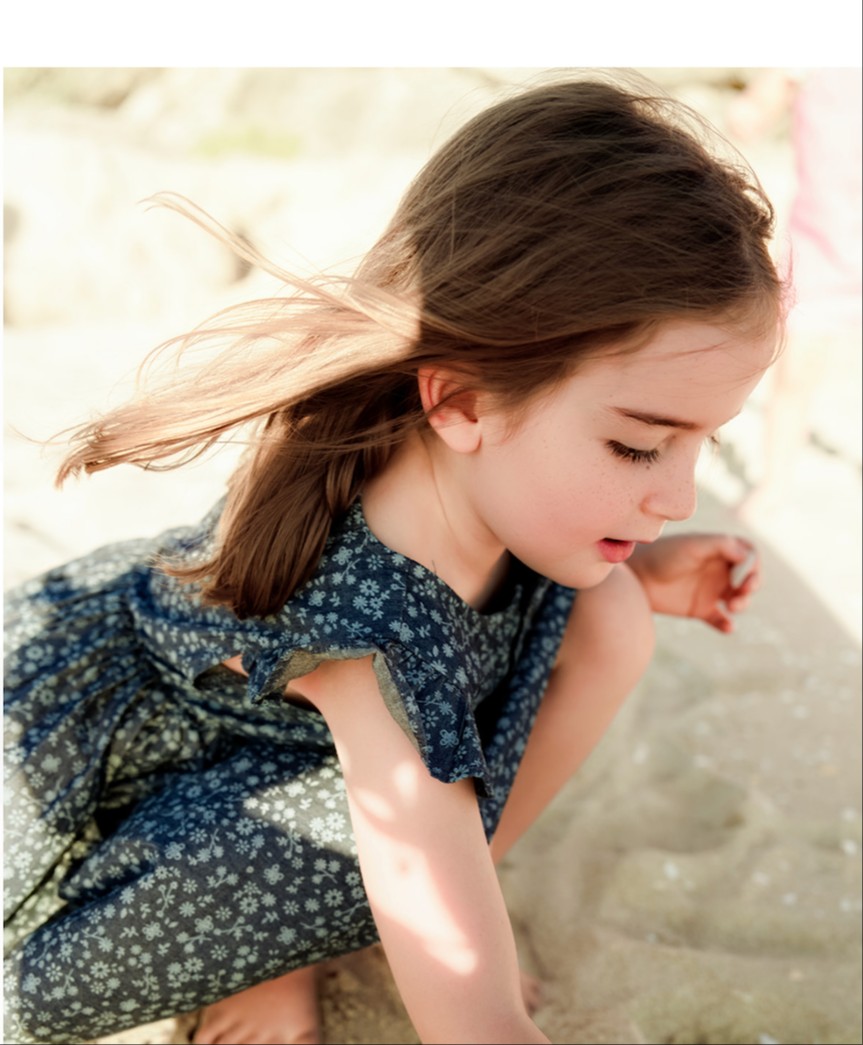 A young girl about 4 years old wearing a dark blue dress, bent down on the sand. Her head is down and we can see half of her face. She has long dark brown hair
