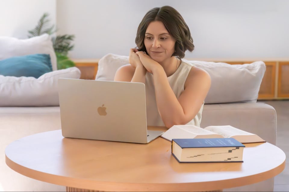 Ash Mastropasqua, homeopath of The Sacred Return. Wearing light, natural / neutral tones made of linen material. Sitting at a desk in front of her computer and homeopathic books on the desk. She having a follow up consultation with a patient via Telehealth