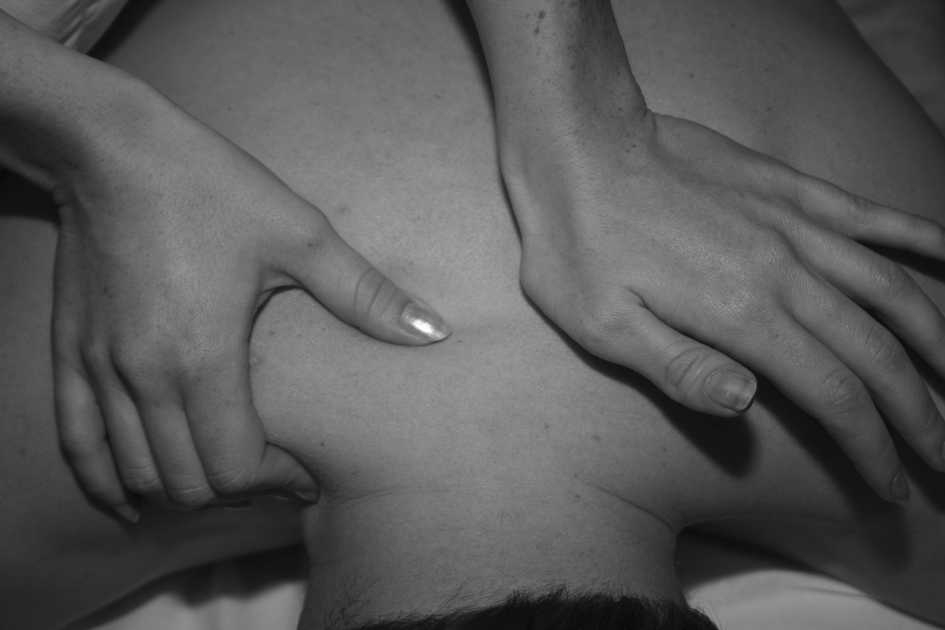 Black and white photos of a female's hands with thumb nails visible, massaging a man's upper shoulders, the therapists right hand is gripping and compressing the man's right upper trapezius muscle and the therapists' left hand is applying pressure through the base of the palm around the upper rhomboid region, with the therapists' fingers slightly lifted and flexed.