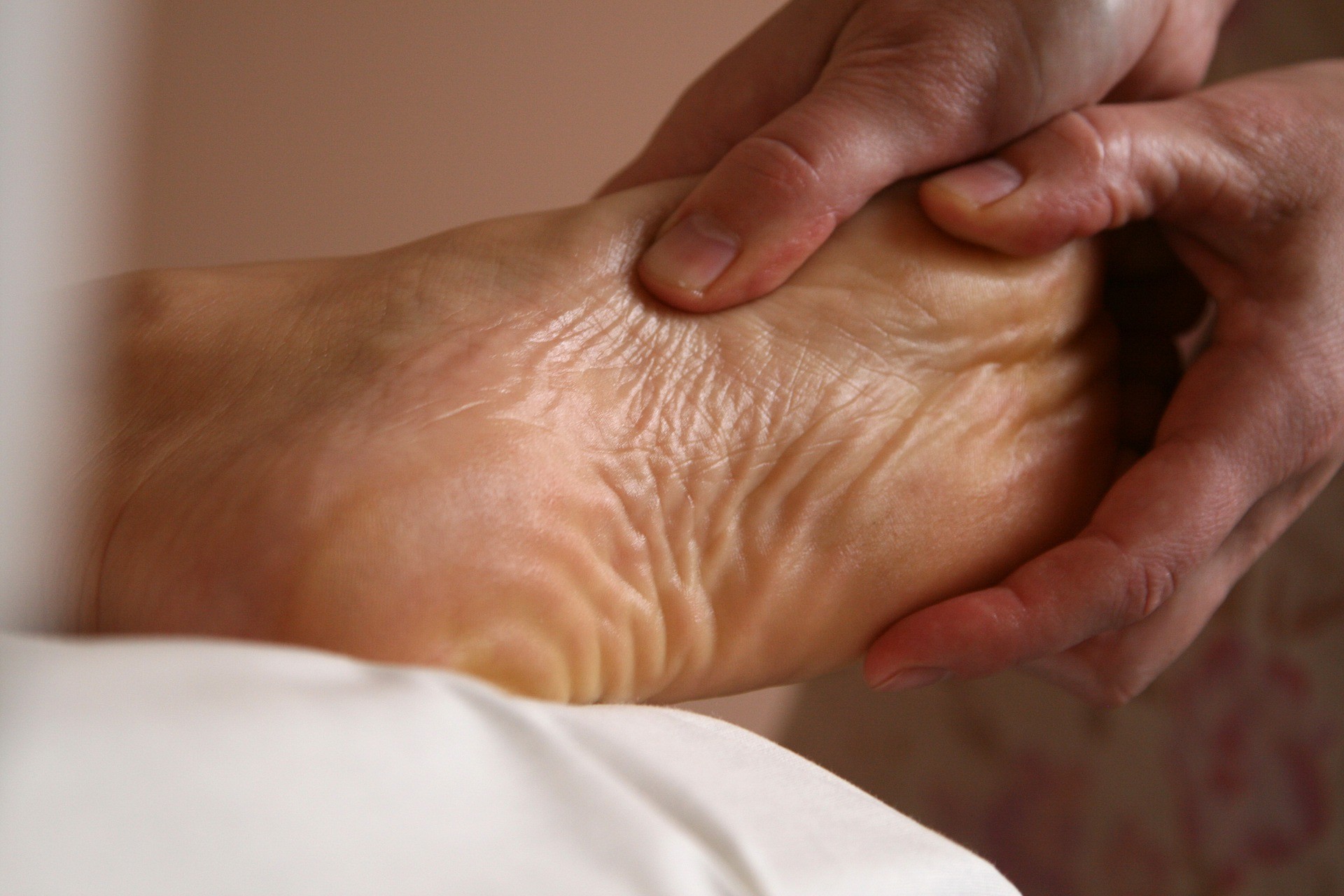 Photo showing thumbs and hands holding and massaging a foot on a client who has their foot elevated on a white pillow, the sole of their foot is crinkling with the massage treatment moving the flesh. The photo is set against a warm pink background and also shows a small part of the therapists' clothes in the background.