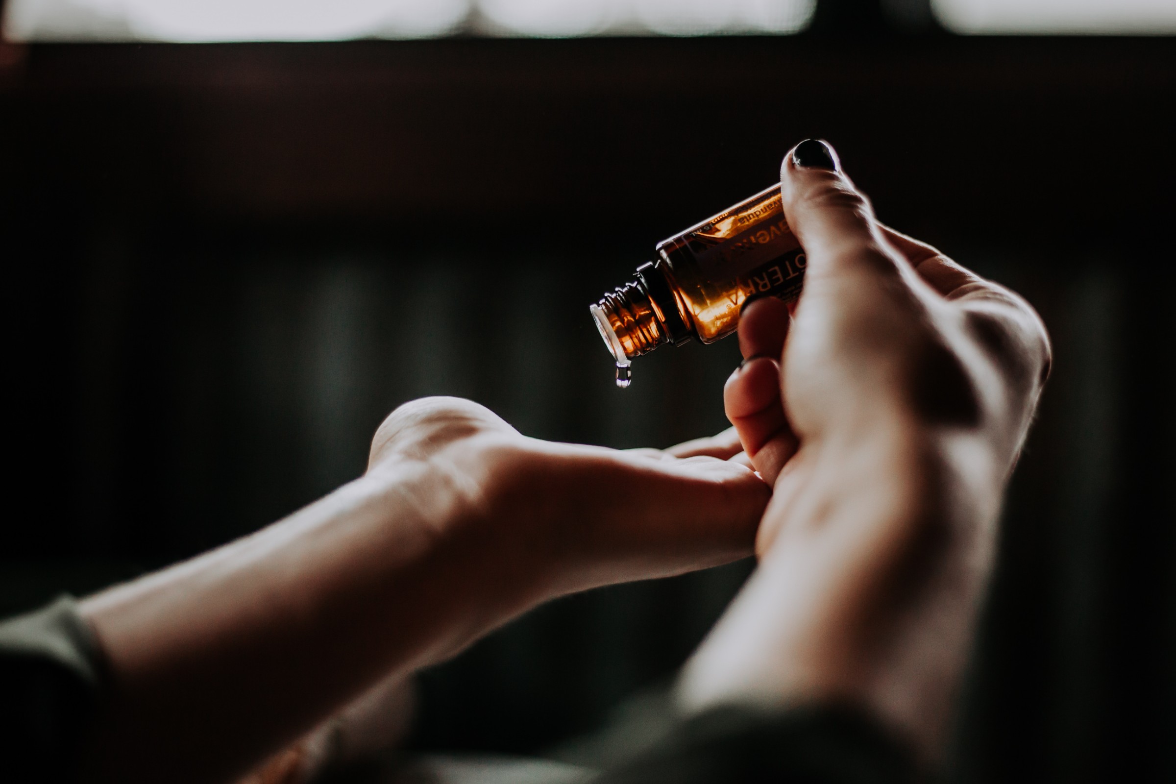 Pouring drops of quality essential oils onto the palm of a hand, you can see one oil drop happening on Caucasian hands against a black background that has a light window in the top upper area of the photo.