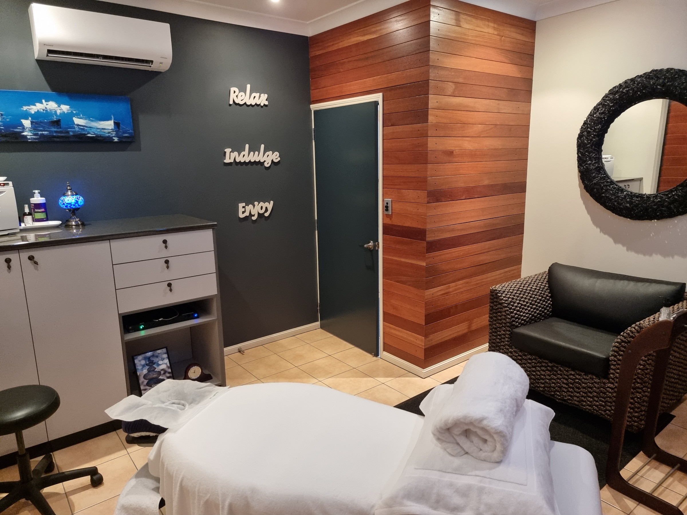 Focus On Movement's massage room, with an electric massage table set up with white linen and a rolled towel on top of a cushion. Comfy black leather chair with wicker sides and a large black round mirror. Stained wood paneling on 2 walls, with a dark blue entry door. On a charcoal feature wall, there are 3 white wooden words hanging up saying "Relax" and "Indulge" and "Enjoy, plus the air-conditioner is visible and a bright blue painting with boats. Grey cabinetry with drawers, a music system and picture frame on an internal open display shelf with a hot towel cabinet and lotions on the top of the cubpoard.