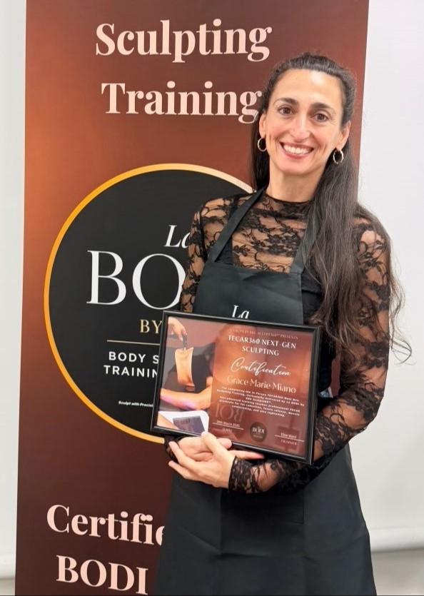 Grace Miano, TECAR360 bodysculptor, nutritionist and women's medicine massage therapist, wearing a black lace long-sleeve top and a clean and crisp black apron, stands smiling and holding a certificate that reads "TECAR360 next-gen sculpting", in front of a large standing sign that reads the same.
