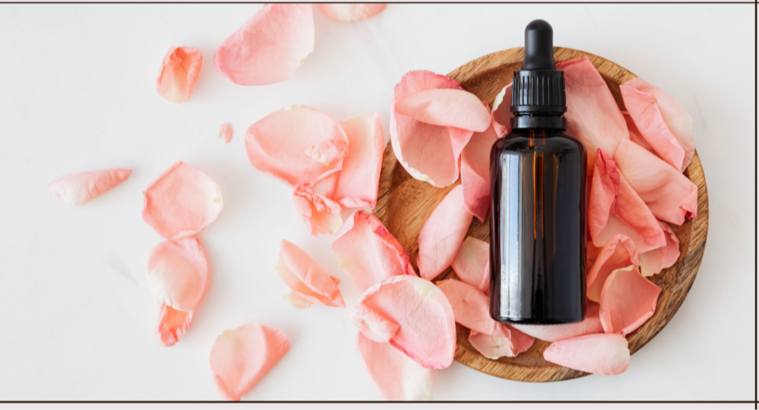 A homeopathic amber dropper bottle with black lid resting upon a wooden bowl that has pink flower petals scattered onto of it and next to the bowl with a white background