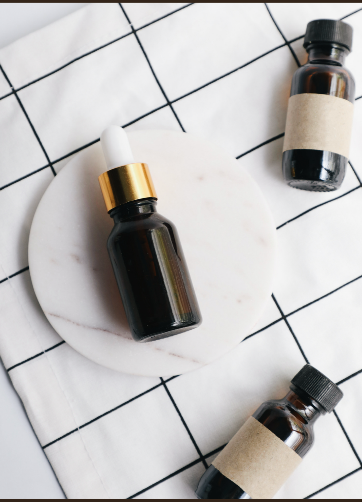 White tiled surface with black grout lines with one full homeopathic amber dropper bottle with gold neck and white lid, surrounded by 2 other smaller amber dropper bottles, one with a blank label across the middle